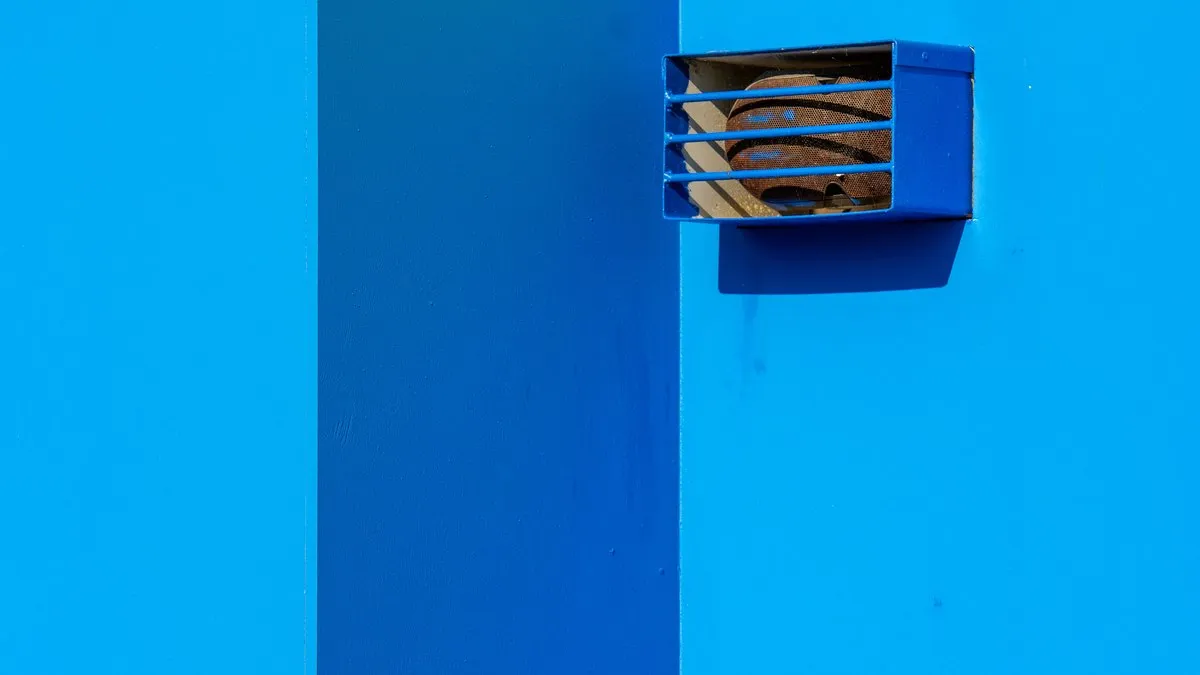 A minimalist shot of a blue wall with rusted vent on sunny day highlighting geometric simplicity.