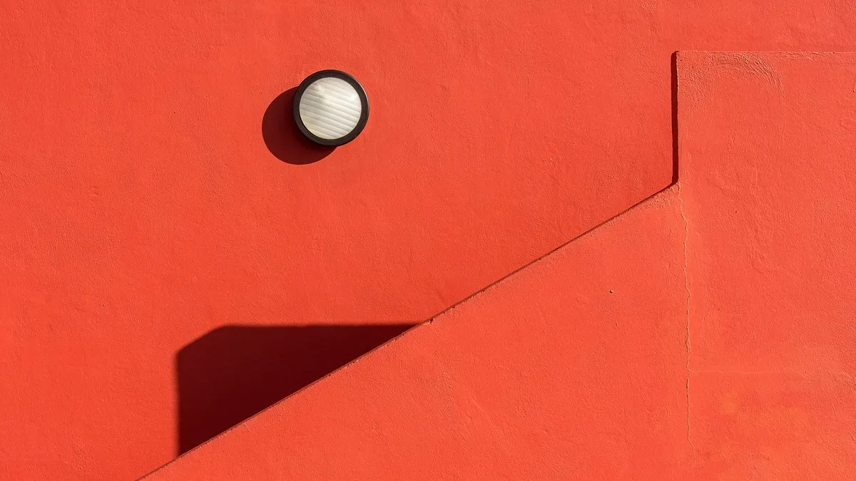 A minimalist red wall featuring a round lamp casting a distinct shadow.