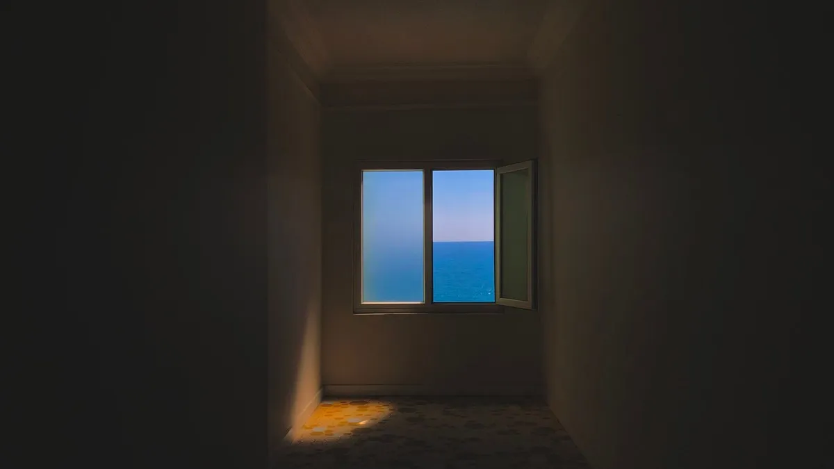 Dark room with an open window showcasing a blue sea horizon.