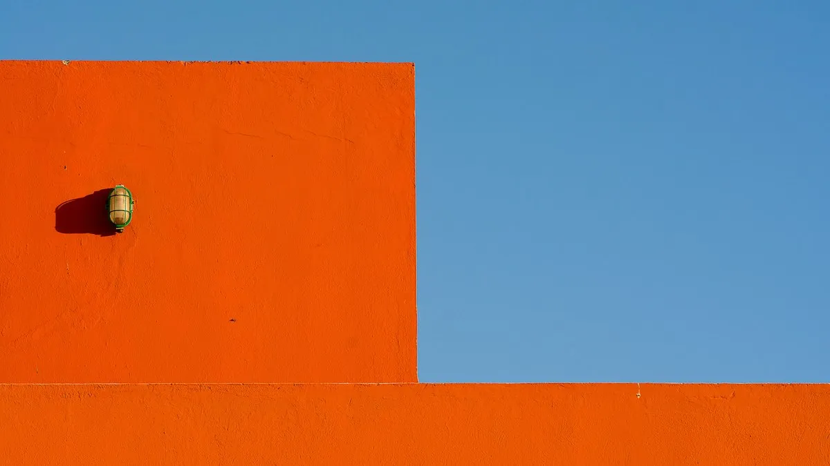 A striking minimalist image of an orange wall featuring a lone lamp against a clear blue sky.