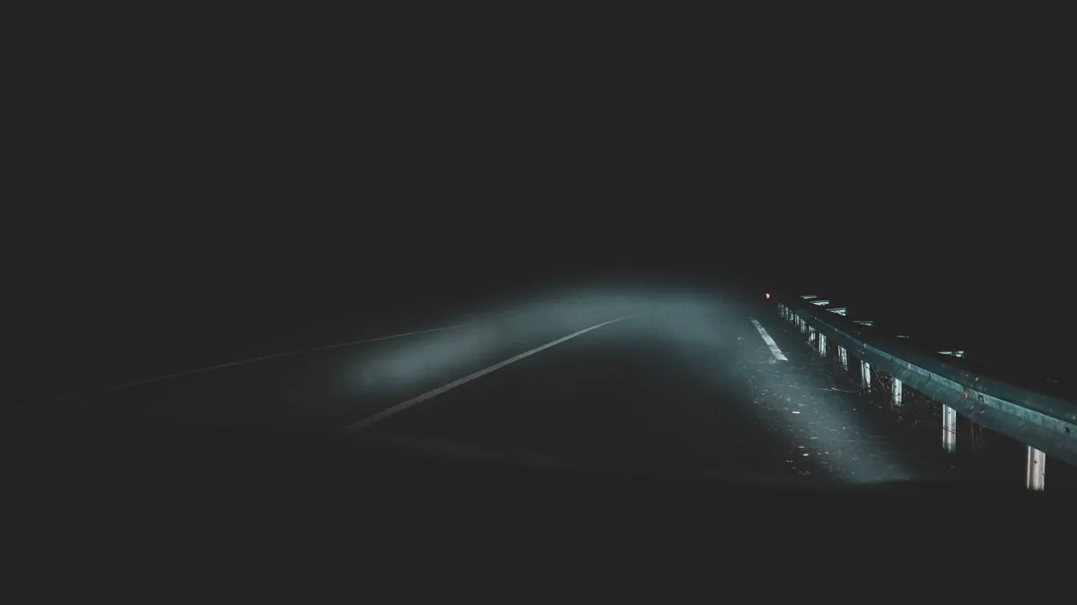 Eerie night road with fog and visible guardrail, illuminated by car headlights, creating a mysterious atmosphere.