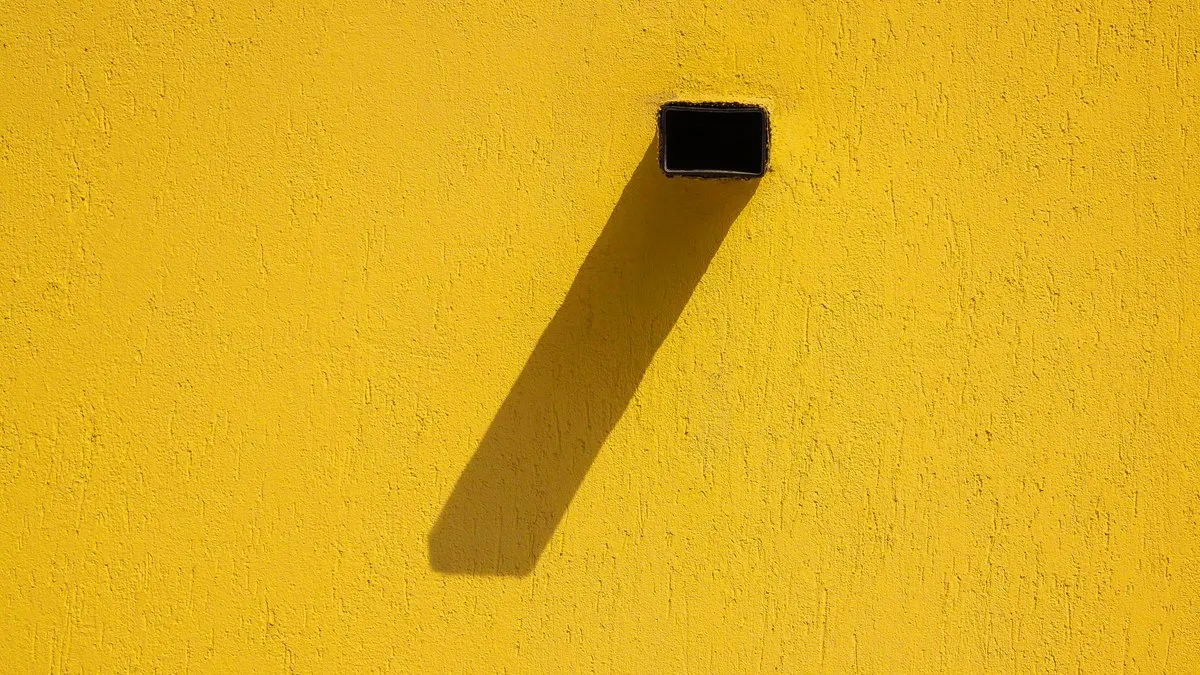 A minimalist composition of a yellow wall with an extended shadow from a protruding pipe.