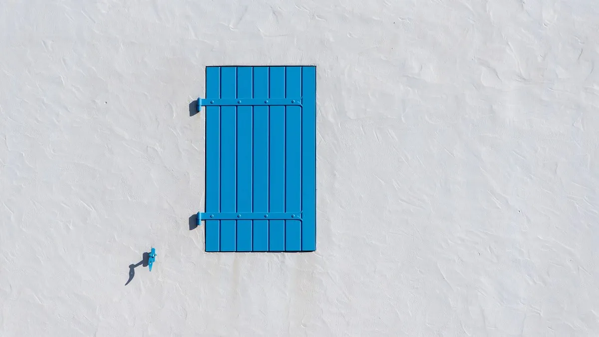 Minimalist photo of a blue shutter on a white wall, showcasing simplicity and contrast.